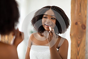 Beauty portrait of pretty young black woman applying lipstick and looking at mirror indoors. Organic makeup products