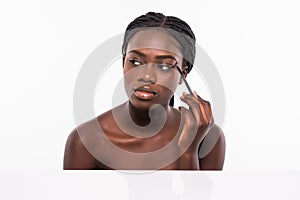 Beauty portrait of a smiling beautiful half naked african woman applying make-up with a brush and looking at camera isolated over