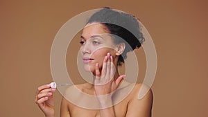 Beauty portrait young African American woman applying skincare serum on face, rubbing gently with fingers. Black female