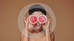 Beauty portrait of young African American woman having fun, covering eyes with halves of grapefruit. Black female on