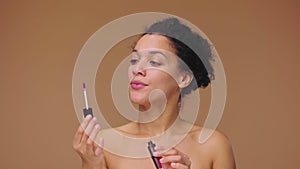 Beauty portrait of young African American woman unrolling lip gloss, examines brush, twirls it back. Black female on