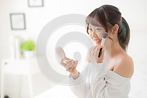 Beauty portrait young asian woman smiling with face looking mirror applying makeup with brush cheek in the bedroom
