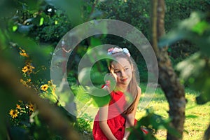 Beauty Girl Outdoors. Beautiful Teenage Model girl in Red Dress is sitting on the garden in Sun Light. Blowing Long Hai