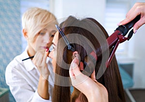 Beauty salon specialists doing professional makeup and hairstyle using curling iron for young woman