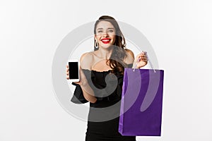 Beauty and shopping concept. Beautiful and stylish woman showing mobile phone screen and bag, buying online, standing