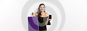 Beauty and shopping concept. Beautiful and stylish woman showing mobile phone screen and bag, buying online, standing