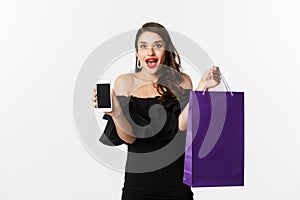 Beauty and shopping concept. Beautiful and stylish woman showing smartphone screen and bag, buying online, standing over