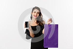 Beauty and shopping concept. Beautiful and stylish woman winking, showing smartphone screen and bag, buying online