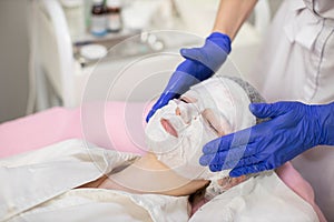 Beauty and skin treatment concept. Young beautiful woman with white moisturizing fabric mask on her face. Hands of