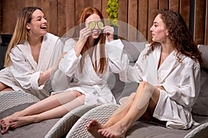 Beauty Spa Day. Cheerful Caucasian Women In Bathrobes Holding Cucumber Slices For Eyes, Going To Do Face Mask Treatment