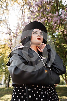 Beauty sunny portrait chic woman in black stylish dress in fashionable hat in vintage leather jacket in park on sunny bright day.