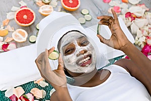 Beauty treatment. Top view of beautiful young African woman lying with pieces of cucumber on her face in spa