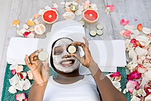 Beauty treatment. Top view of beautiful young African woman lying with pieces of cucumber on her face in spa