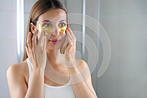 Beauty woman applying golden anti-aging under-eye mask looking herself in the mirror in the bathroom. Skin care girl touch patches