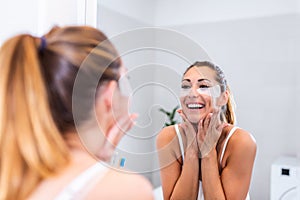 Beauty woman applying under-eye mask looking herself in the mirror in the bathroom. Skin care girl touch patches of fabric mask