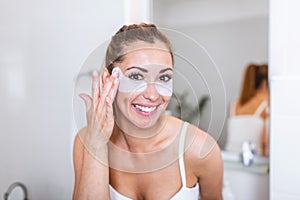Beauty woman applying under-eye mask looking herself in the mirror in the bathroom. Skin care girl touch patches of fabric mask