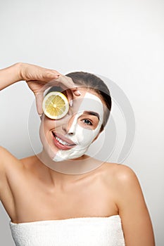 Beauty Woman Face Mask. Portrait Of Beautiful Girl With Cosmetic White Mask On Facial Skin Holding Slices of Fresh Lemon
