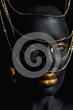 Beauty woman painted in black skin color body art, gold chain in his hands and around his neck. Gold makeup lips eyelids,