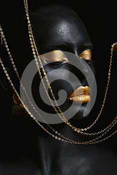 Beauty woman painted in black skin color body art, gold chain in his hands and around his neck. Gold makeup lips eyelids,