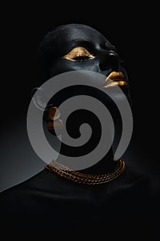 Beauty woman painted in black skin color body art, gold chain in his hands and around his neck. Gold makeup lips eyelids,