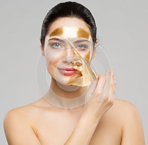 Beauty Woman peeling off Golden Facial Mask. Smooth Skin Model taking off Gold Purifying Face Film Mask over Gray. Women cleansing photo