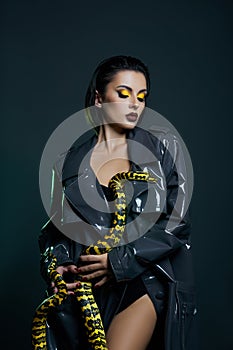 Beauty woman python yellow snake around her neck on latex shiny raincoat. Yellow snake on the shoulders of girl. Beauty yellow eye