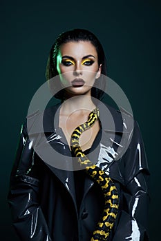 Beauty woman python yellow snake around her neck on latex shiny raincoat. Yellow snake on the shoulders of girl. Beauty yellow eye