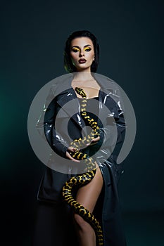 Beauty woman python yellow snake around her neck on latex shiny raincoat. Yellow snake on the shoulders of girl. Beauty yellow eye