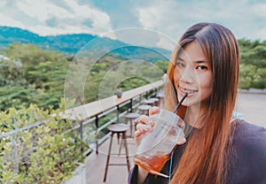 Beauty smiling female is in Cafe with forest and mountain nature background while drinking iced americano coffee photo