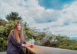 Beauty Young smiling female is sitting in Cafe with forest and mountain nature background while drinking iced americano photo