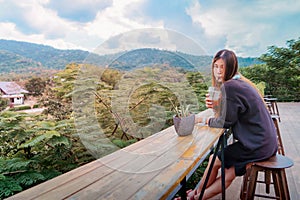 Beauty Asian smiling female is sitting in Cafe with forest and mountain nature background while drinking iced americano photo