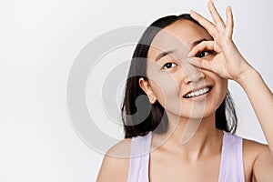 Beauty. Young asian woman with clean glowing skin, showing ok, zero gesture against eye, smiling and gazing away
