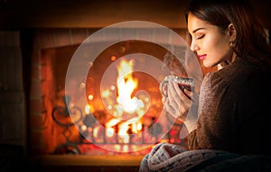 Beauty young Christmas woman sitting near fireplace in dark room at home and drinking hot beverage from a big cup