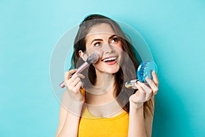 Beauty. Young glamour girl applying make up with pocket mirror and brush, smiling and gazing up at logo, standing