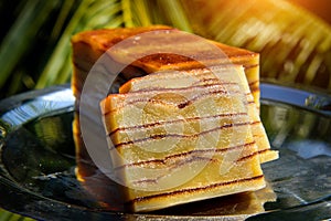 Bebinka on platter in sunlight, blurred leafy background, close-up. Bibik, the national Goan pie, cut into slices. Multi-layer