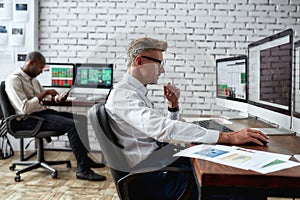 Becoming financially strong. Middle-aged trader sitting by desk in front of computer monitor while working in the office