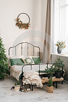 Bed with a beige plaid and green pillows, a Christmas wreath on the wall, a Christmas tree. New Year`s winter home interior decor