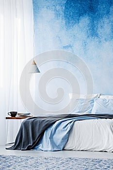 Bed with grey blanket against blue ombre wall in bedroom interior with lamp above table