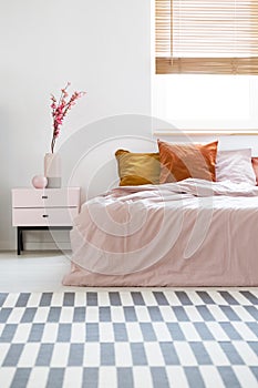 Bed with pink sheets and orange cushions standing in white bedroom interior with carpet, window with wooden blinds and bedside ta