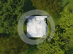 A bed with white linens stands outdoors on the grass in the meadow. White bed in open air surrounded by a landscape in a top view photo