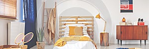 Bed with wooden bedhead, white sheets, ochre cushion and yellow knit blanket