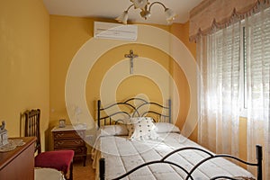 Bedroom with a small bed with a black metal frame, wooden bedside tables and yellow painted walls and a crucifix on the wall