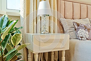 Bedside table, lamp with lampshade, in the interior of a classic bedroom in light pink colors