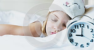 Bedtime. Little girl goes to sleep early evening with alarm clock on 9 p.m., wearing white cat, bunny sleep mask. Soft bed with