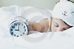 Bedtime. Little girl goes to sleep early evening with alarm clock on 9 p.m., wearing white cat, bunny sleep mask. Soft bed with