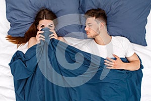 Bedtime. Young couple lying on bed man sleeping woman hiding under blanket playful top view