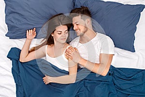 Bedtime. Young couple lying on bed under blanket top view hugging waking up cheerful