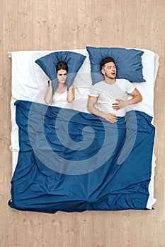 Bedtime. Young man sleeping yawning while woman covering ears with pillow looking at him unhappy under blanket on bed