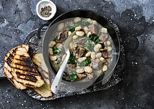Beef, beans, spinach slow cooker stew in a pan on a wooden board on a dark background. Delicious homemade comfort food