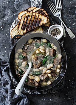 Beef, beans, spinach slow cooker stew in a pan on a wooden board on a dark background. Delicious homemade comfort food